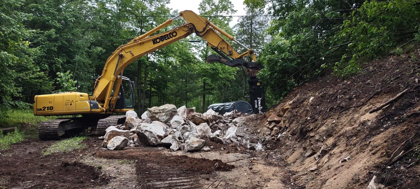 Large Kobelco excavator breaking and clearing boulders along a forested slope during a rural site preparation project, with hydraulic rock breaker attachment in use.