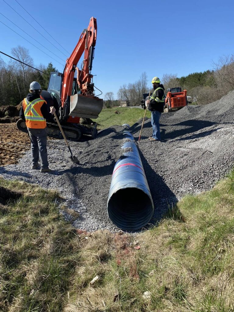 Construction crew installing a culvert pipe during a drainage project in rural Kingston, Ontario. Excavator and workers backfilling with gravel for proper water flow and erosion control.