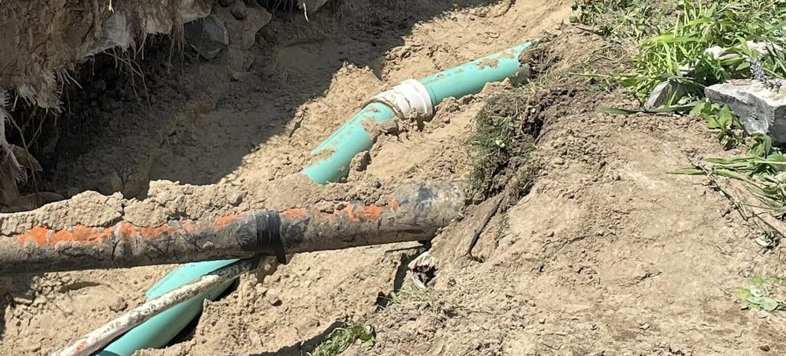 Exposed underground utility trench with damaged green sewer pipe and older corroded water line, surrounded by broken rock and loose soil during an excavation repair project.