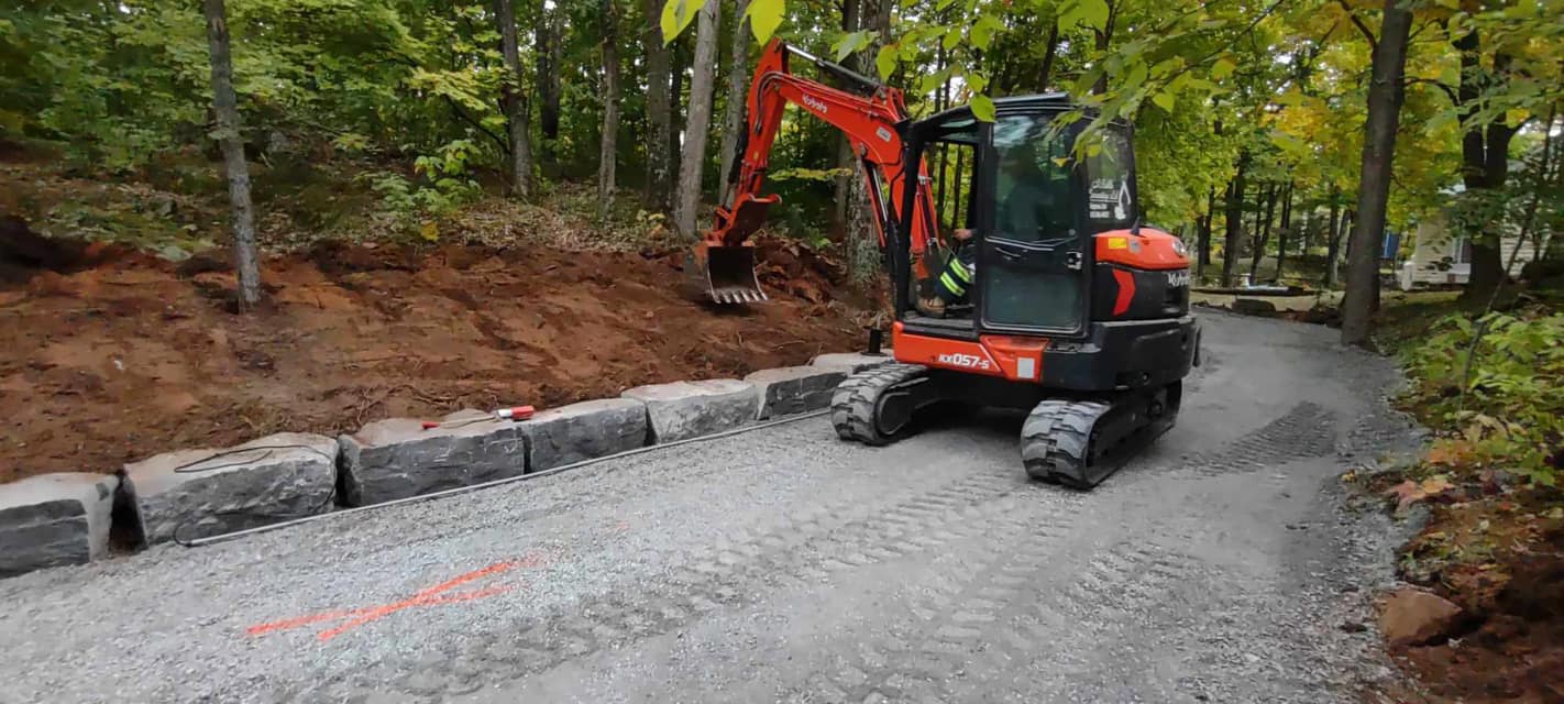 Excavator grading a gravel cottage lane with stone edging, part of road preparation work to ensure proper drainage and a stable surface.