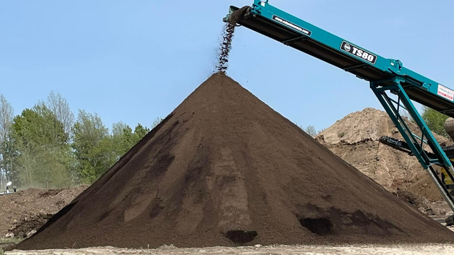 Screened topsoil being piled into a large cone shape by a conveyor, showing the rich texture and colour of good soil prepared for landscaping and gardening projects.