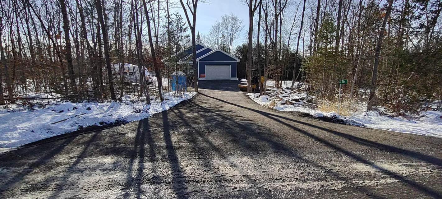 Driveway in the winter with snow on the ground and a blue home in the background.