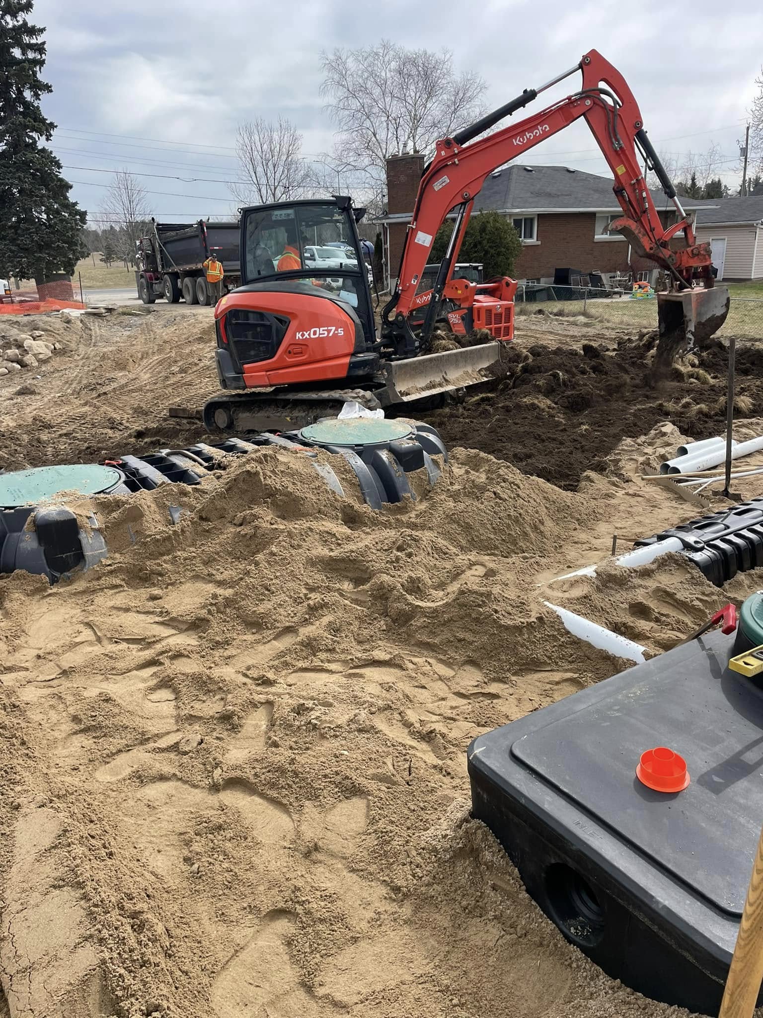 Excavator installing a septic system drain field with plastic chambers and sand backfill at a residential property.