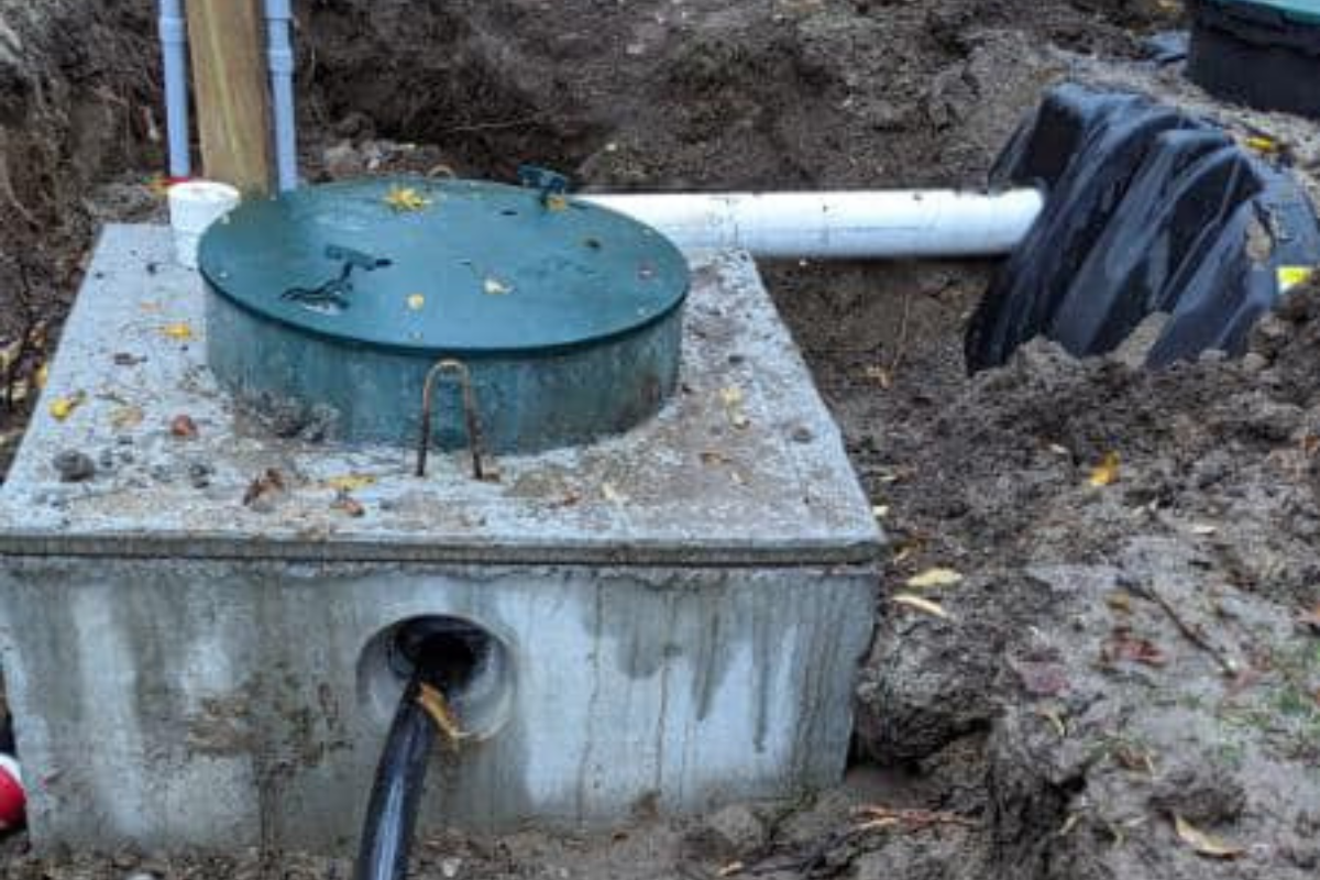 Concrete septic tank set in place with inlet and outlet pipes connected. Electrical conduit and risers positioned for access and monitoring.