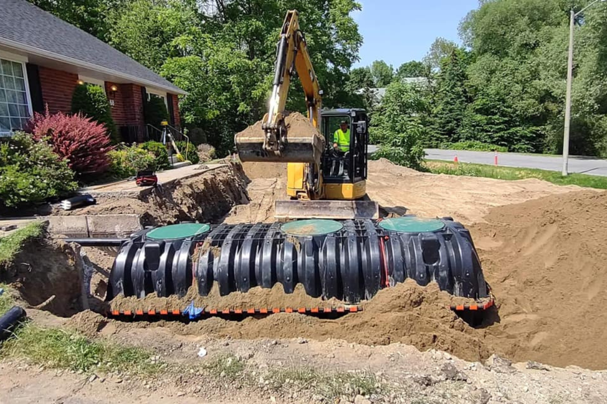 Excavator backfilling around a new plastic septic tank beside a residential home.