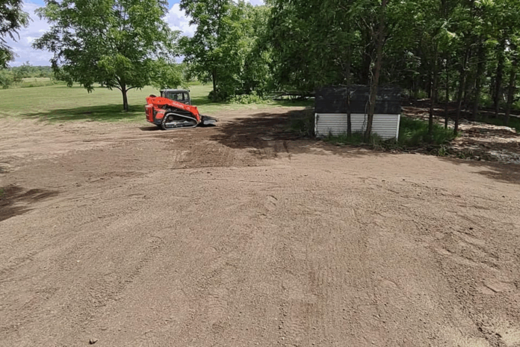 Finished grading over a septic installation site, with a compact loader in the background near a tree line and utility building.