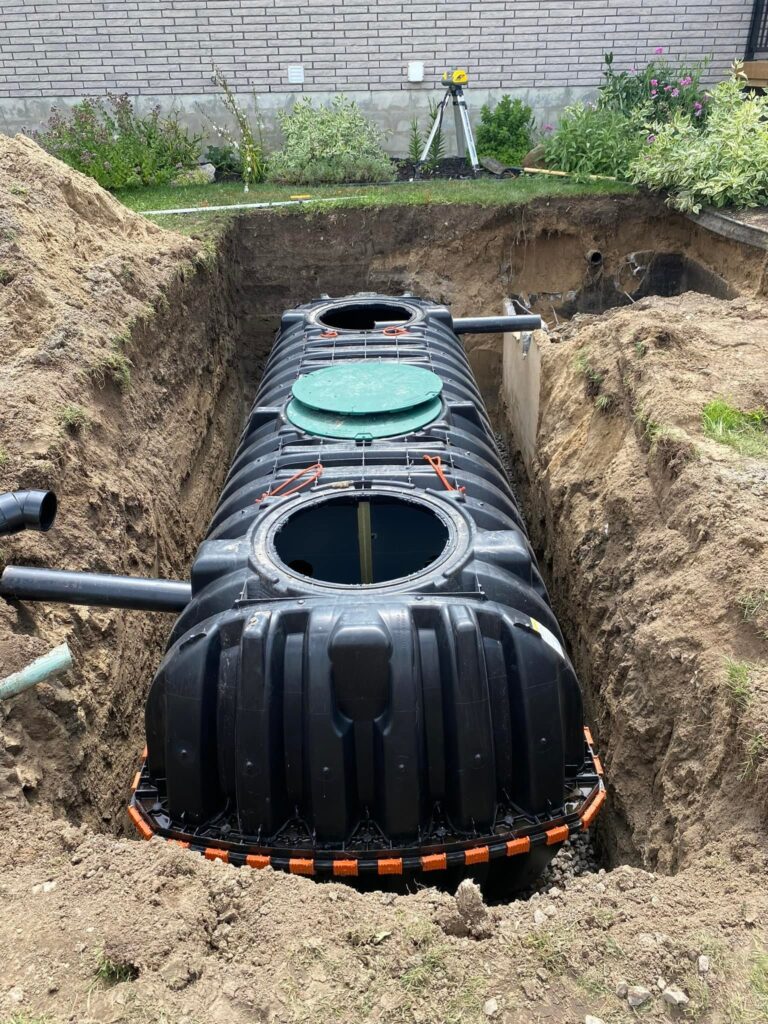 Excavator placing a septic tank with green access lids into a prepared trench during a residential septic system installation.