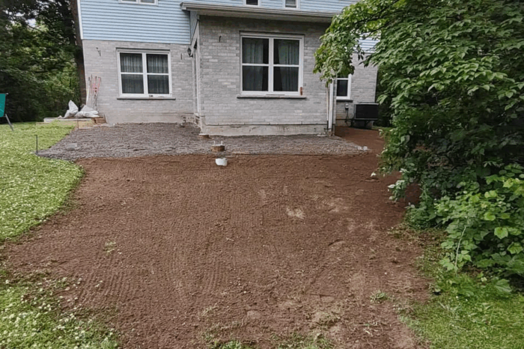 Wide view of gravel pad and topsoil grading work completed behind a two-story home, surrounded by mature trees and lawn. fill to level a yard. Shed & pool site preparation