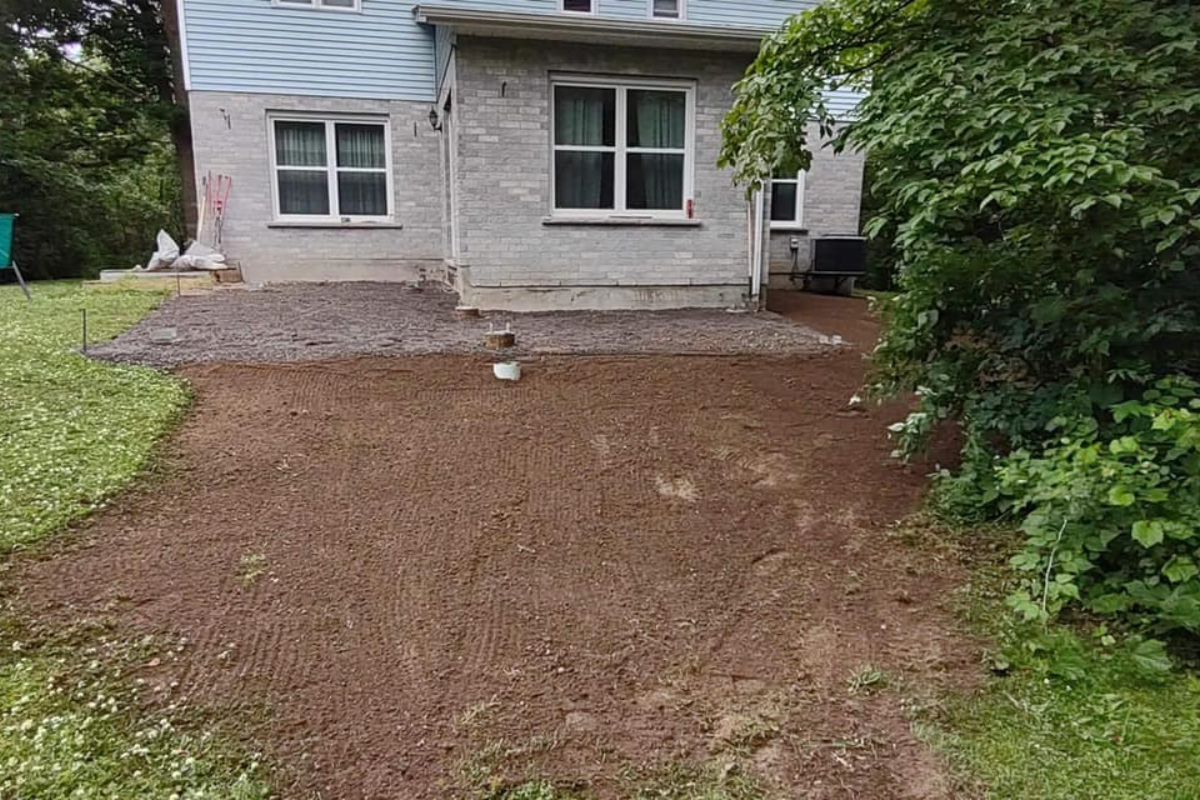 Wide view of gravel pad and topsoil grading work completed behind a two-story home, surrounded by mature trees and lawn.