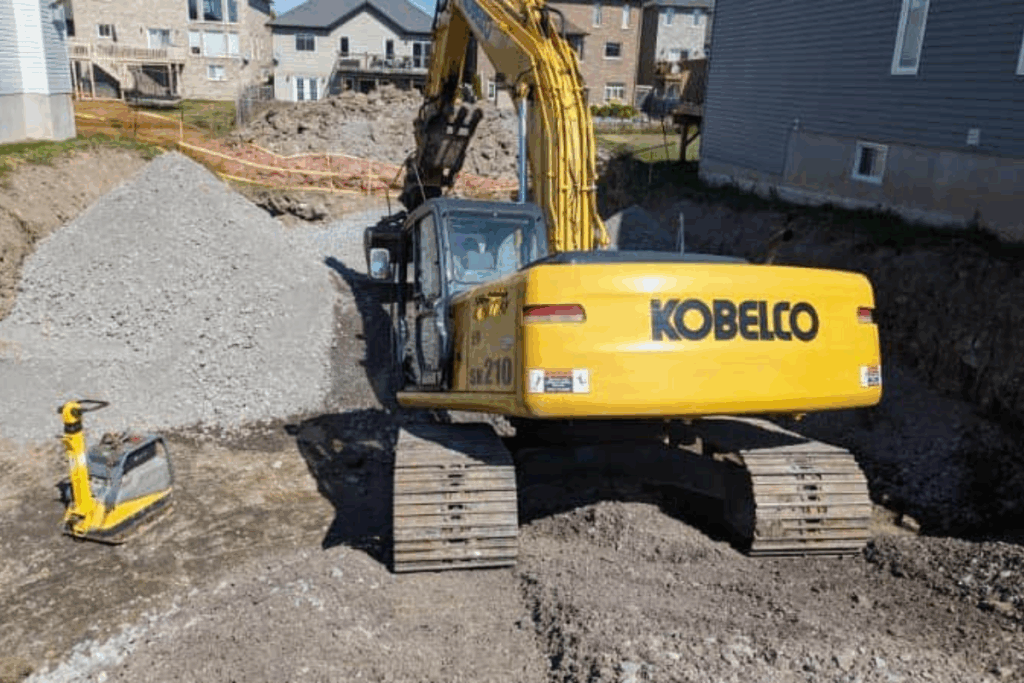 Excavator on a residential property site in Westbrook digging around the foundation to expose soil and gravel for waterproofing and drainage improvement.