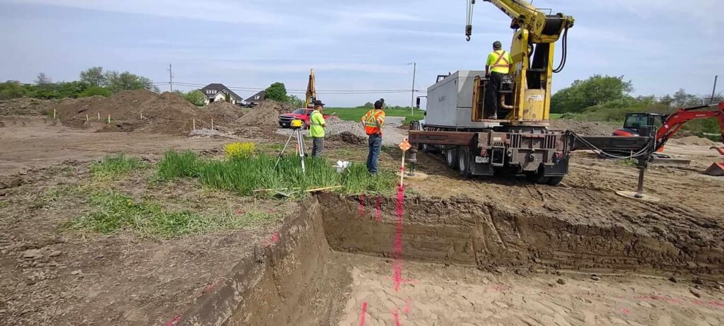 Utility locates marked on soil with workers standing beside an excavator.