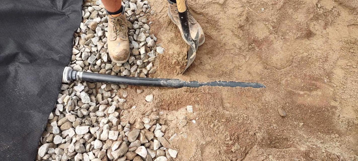 Clay soil and gravel underneath a drainage pipe with the boots of a construction worker standing on top.